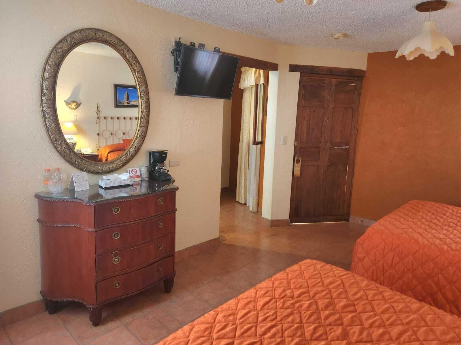 A hotel room with a wooden dresser, a large mirror, a flat-screen TV, and an orange quilted bed.