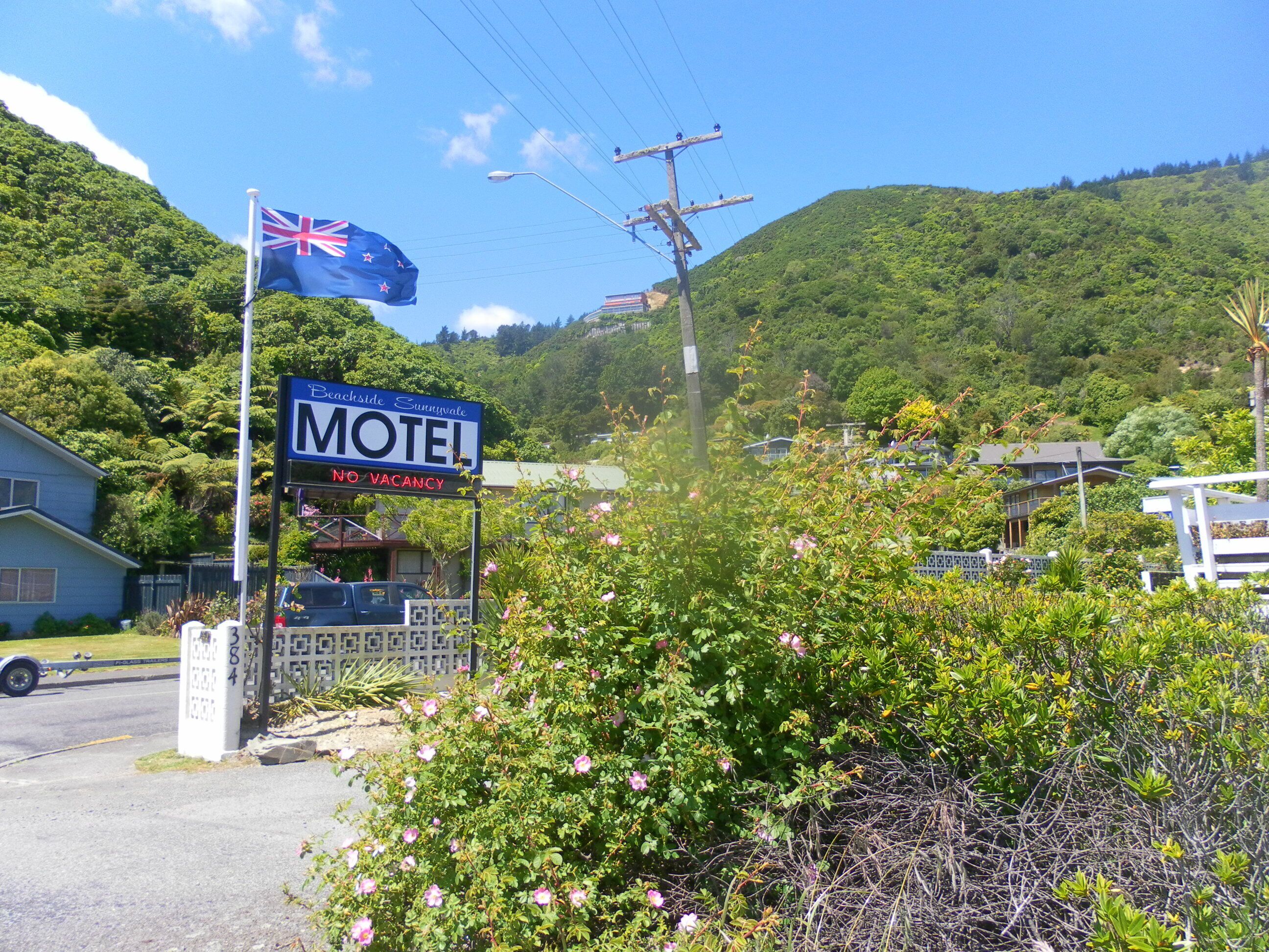 A motel sign with a flag, surrounded by greenery and a hill in the background.
