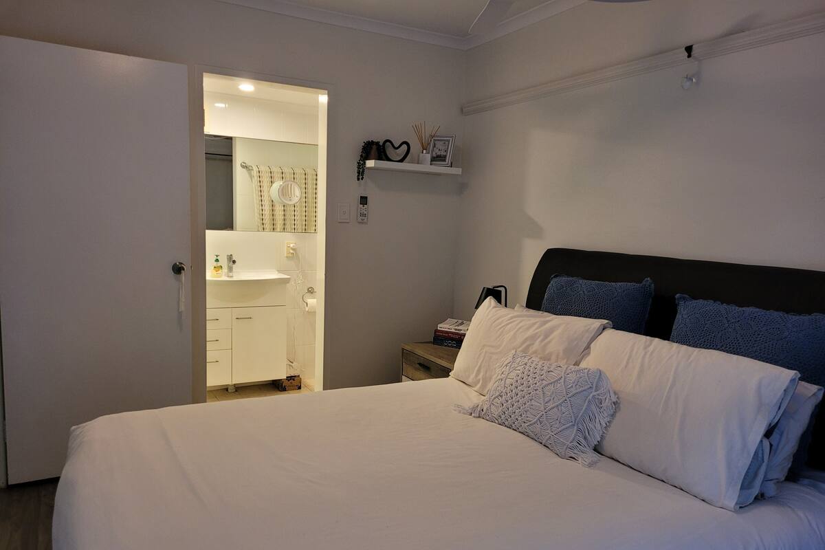 Une chambre à coucher avec un grand lit, une table de chevet, une étagère ornée d’objets décoratifs et une salle de bain visible à travers une porte ouverte.