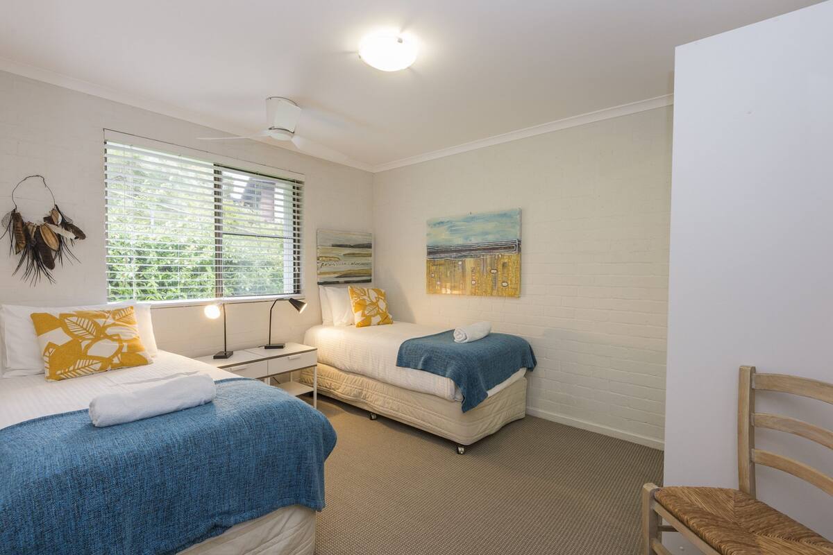 Une chambre avec deux lits, un bureau, une chaise, une fenêtre avec des stores et un tableau au mur.