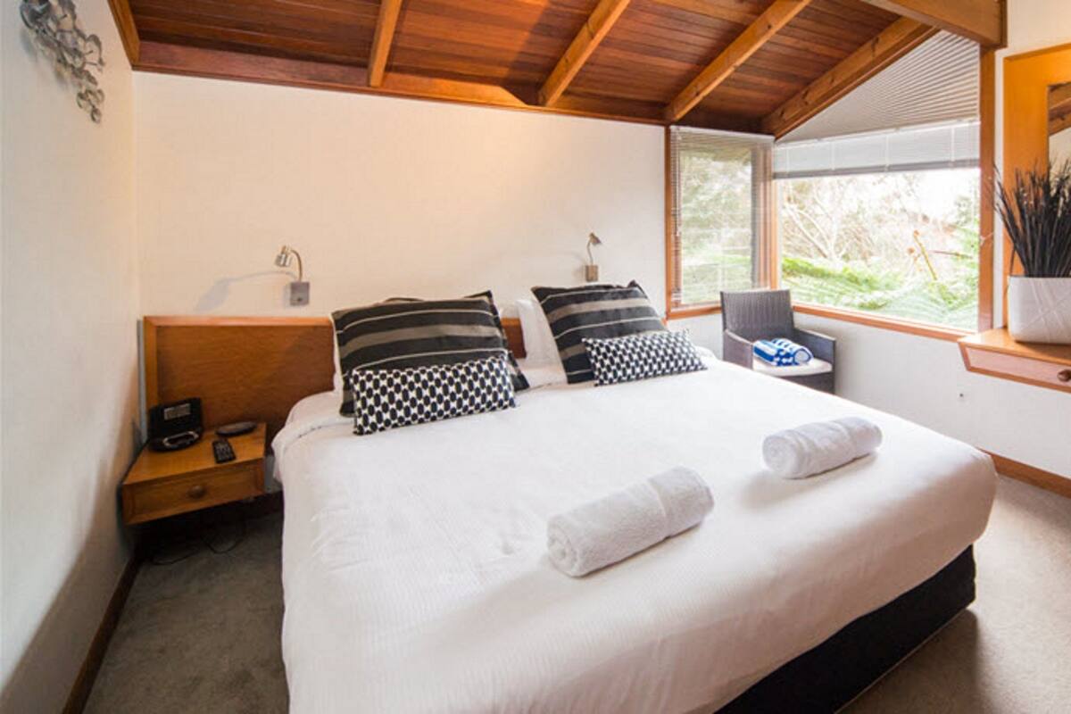 Une chambre avec un grand lit, une table de chevet avec une lampe, une chaise, une petite table avec une plante et une fenêtre donnant sur un paysage verdoyant.