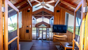 A spacious room with a wooden ceiling, a ceiling fan, a television, and large windows offering a view of the outdoors. - The Oasis Apartments and Treetop Houses (Byron Bay)