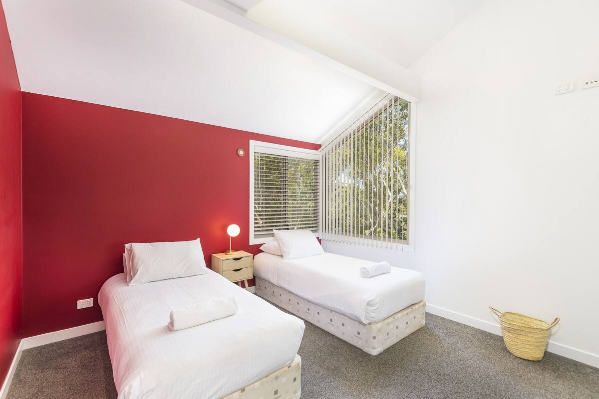 Une chambre avec deux lits, une fenêtre avec des stores, une table de chevet avec une lampe et un petit panier en osier.