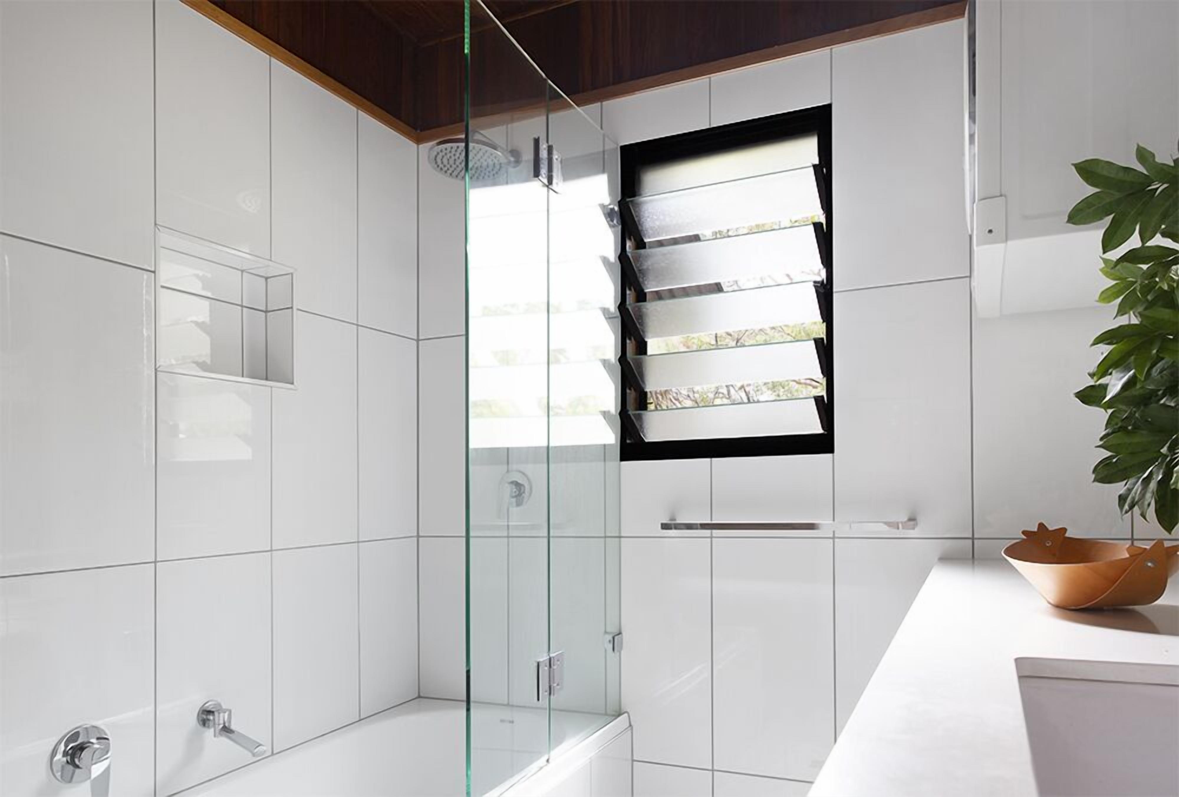 A modern bathroom with white tiles, a glass shower enclosure, and a small window.