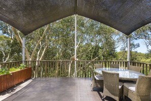A covered outdoor seating area with a table and chairs, surrounded by a wooden railing and a view of trees.