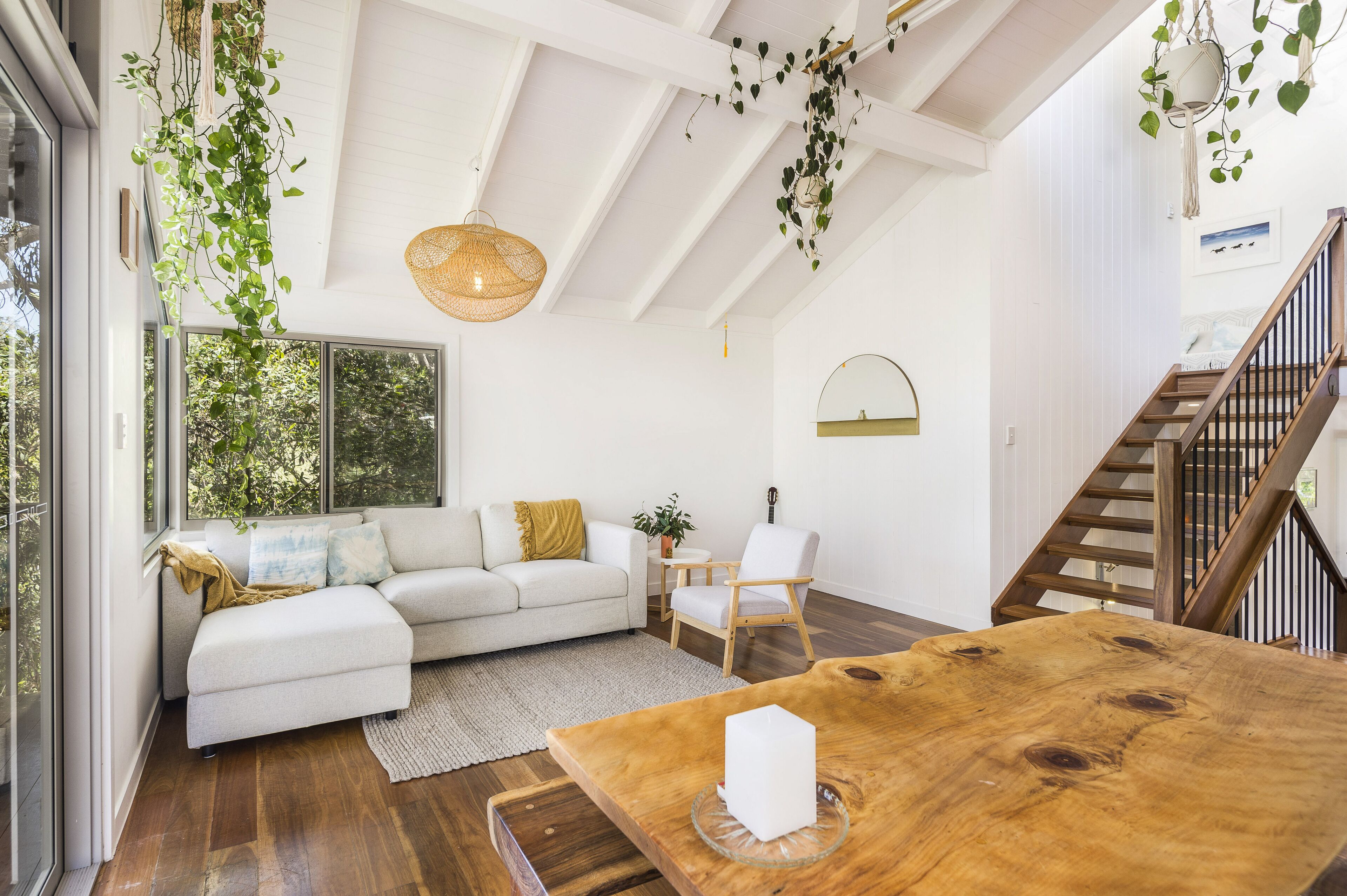 A spacious living area with a large wooden table, a white sofa, and a wooden chair. There is a staircase with wooden railings and a hanging plant.