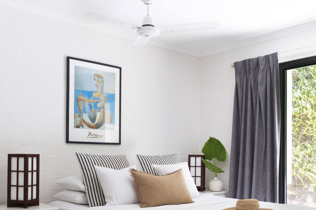 Une chambre à coucher bien rangée, avec un lit, des oreillers, une table de chevet, un tableau encadré, une plante et une fenêtre avec des rideaux.