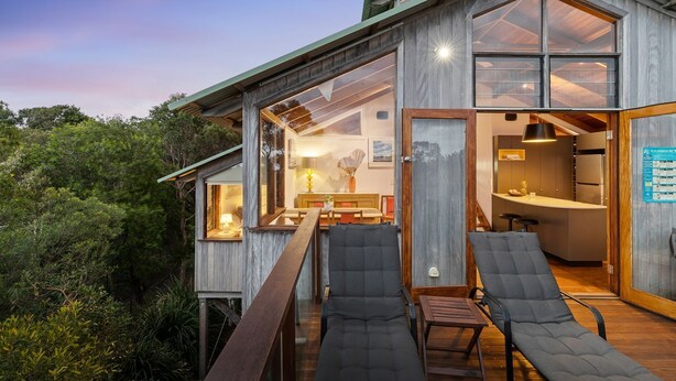 A modern wooden house with a deck, large windows, and a kitchen area visible through the open door. - The Oasis Apartments and Treetop Houses (Byron Bay)