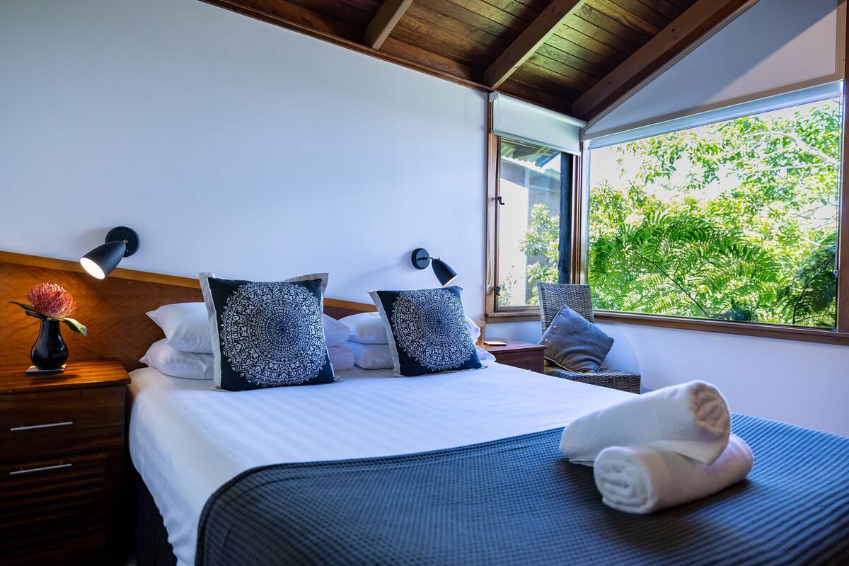 Un lit bien fait, avec du linge de lit blanc et des coussins à motifs, une lampe de chevet et une vue sur la verdure par la fenêtre.