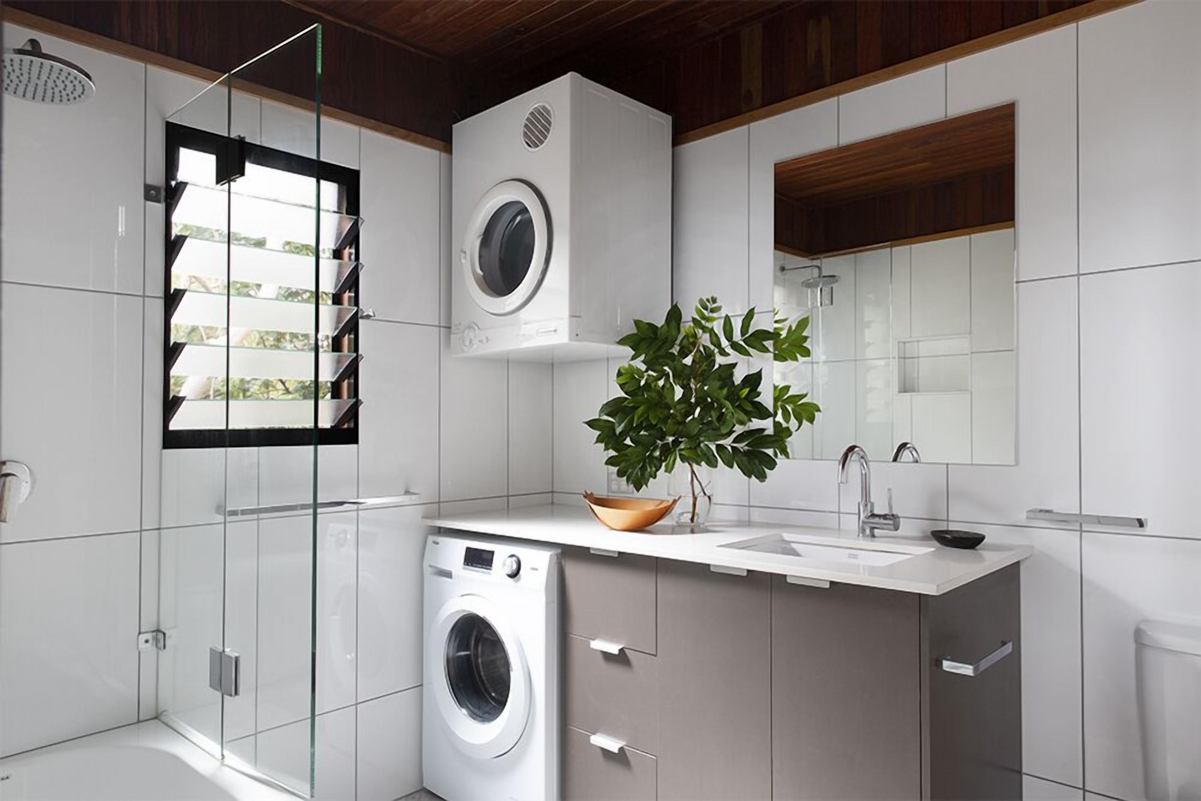 A modern bathroom with a glass shower enclosure, a built-in washing machine, a sink with a mirror, and a potted plant.
