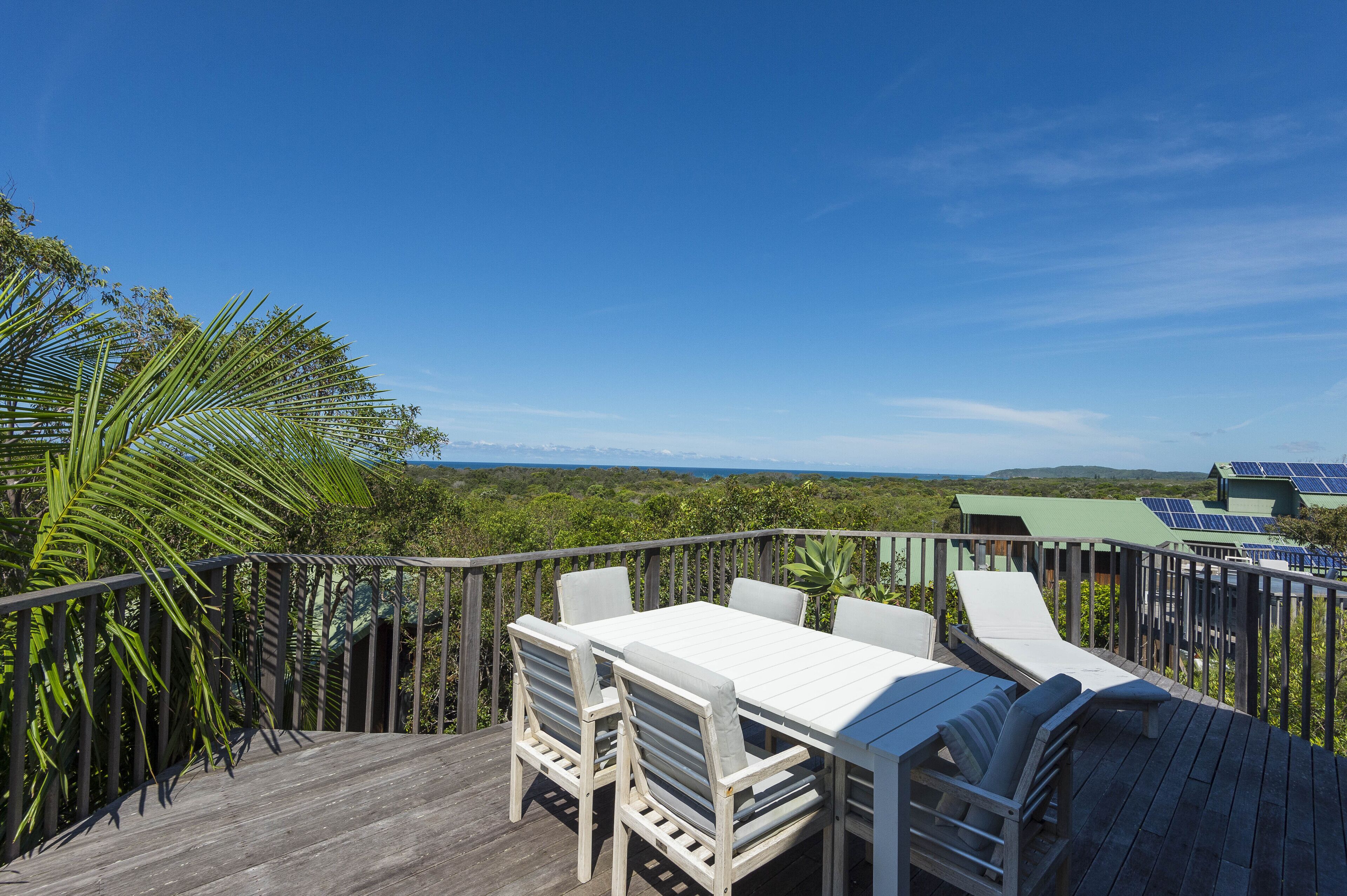 Une terrasse en bois avec une table et des chaises blanches, entourée d’une végétation luxuriante et sous un ciel bleu azur.