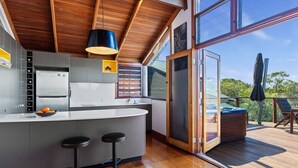 A modern kitchen with a central island, bar stools, and a large window offering a view of the outdoors. - The Oasis Apartments and Treetop Houses (Byron Bay)