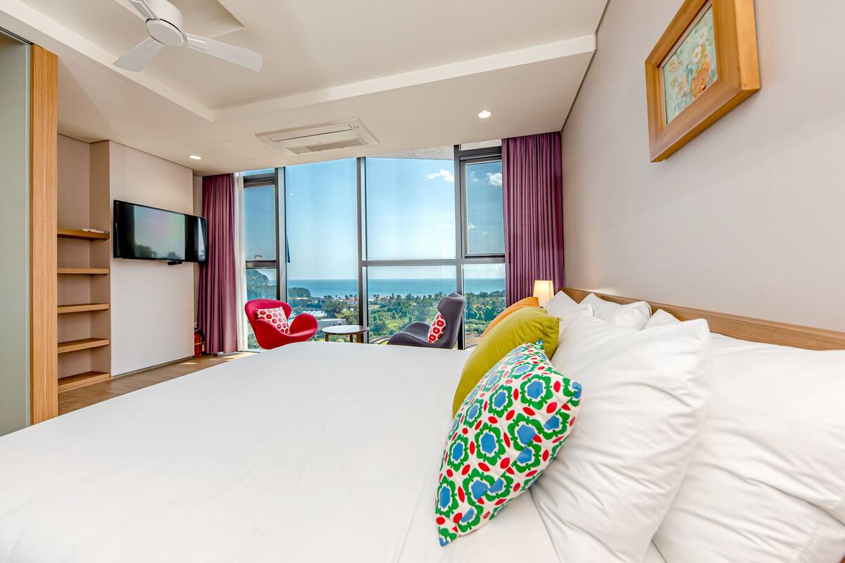 A hotel room with a large bed, a TV, a red chair, and a view of the city.