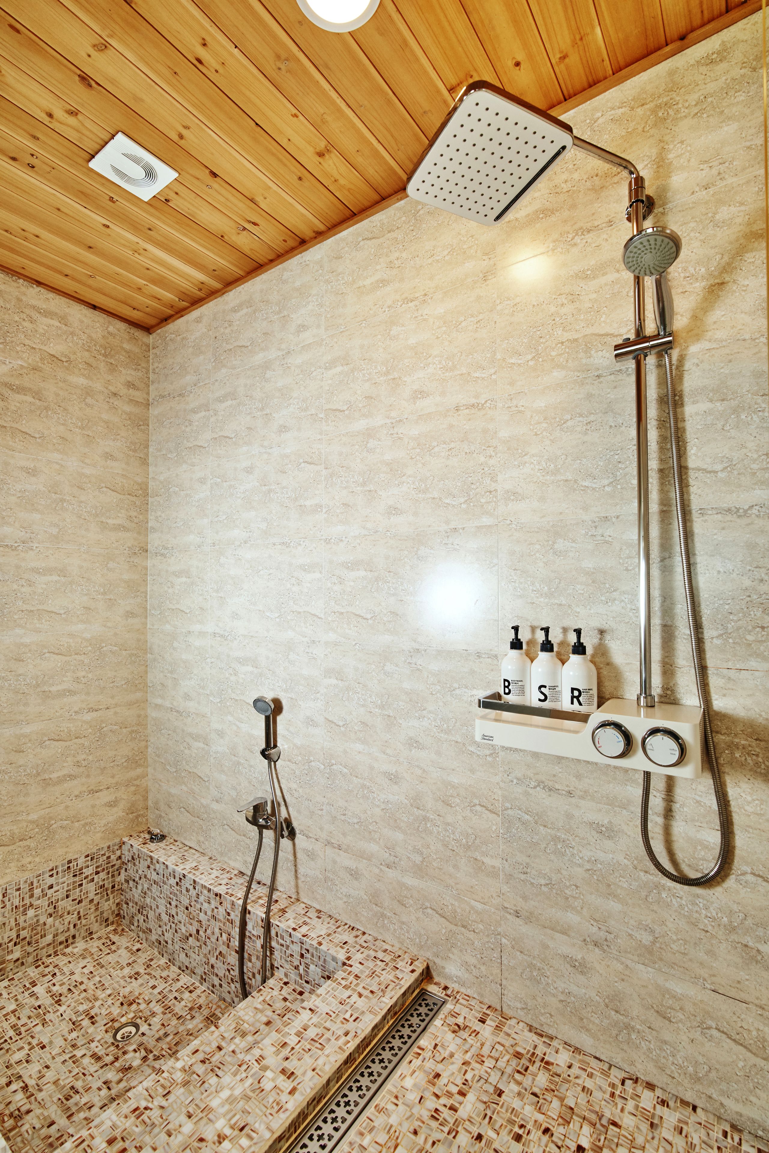 A shower area with a wooden ceiling, a tiled floor, and a wall-mounted shelf with bottles.