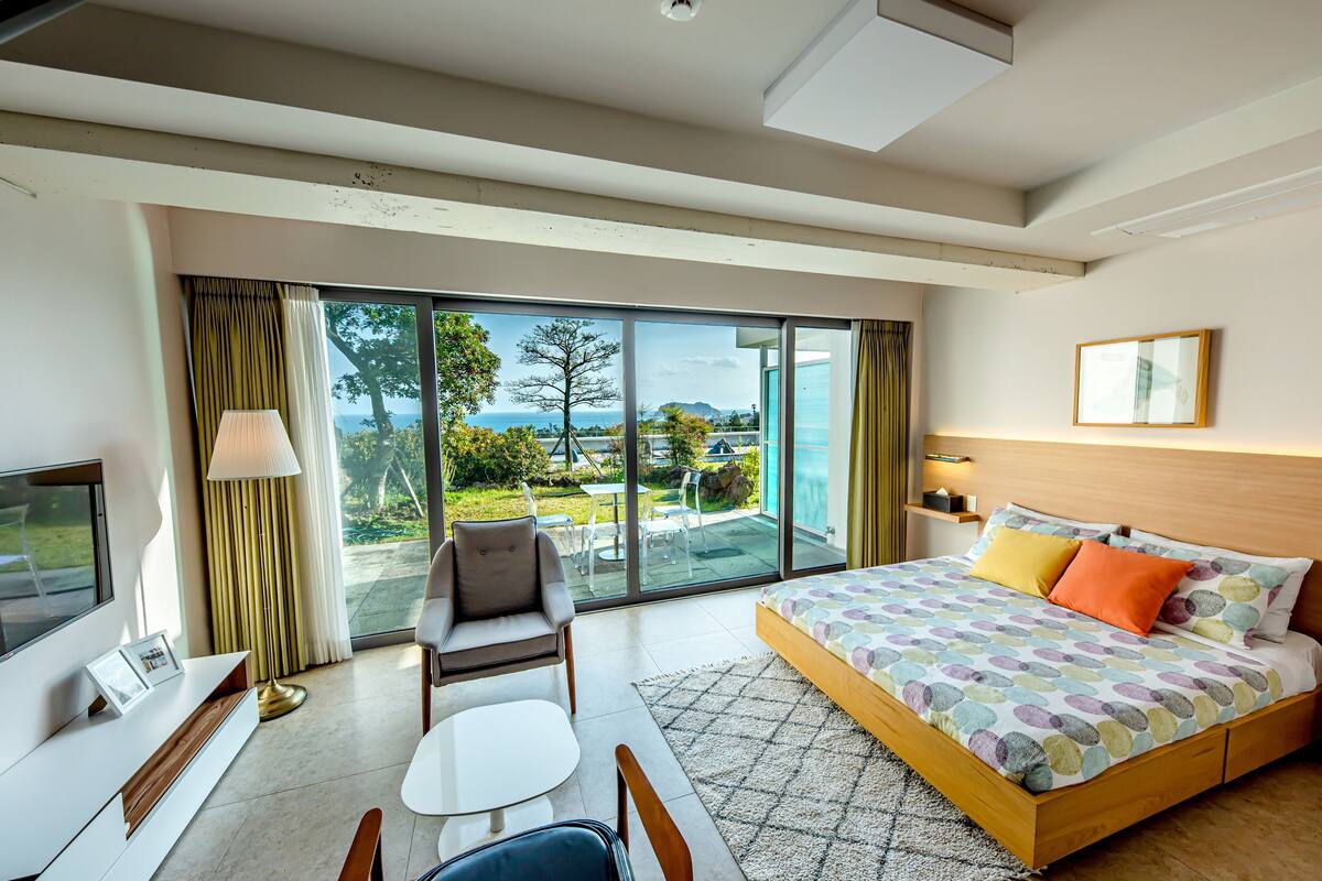 A modern hotel room with a large bed, a TV, a desk, and a balcony with a view of the outdoors.