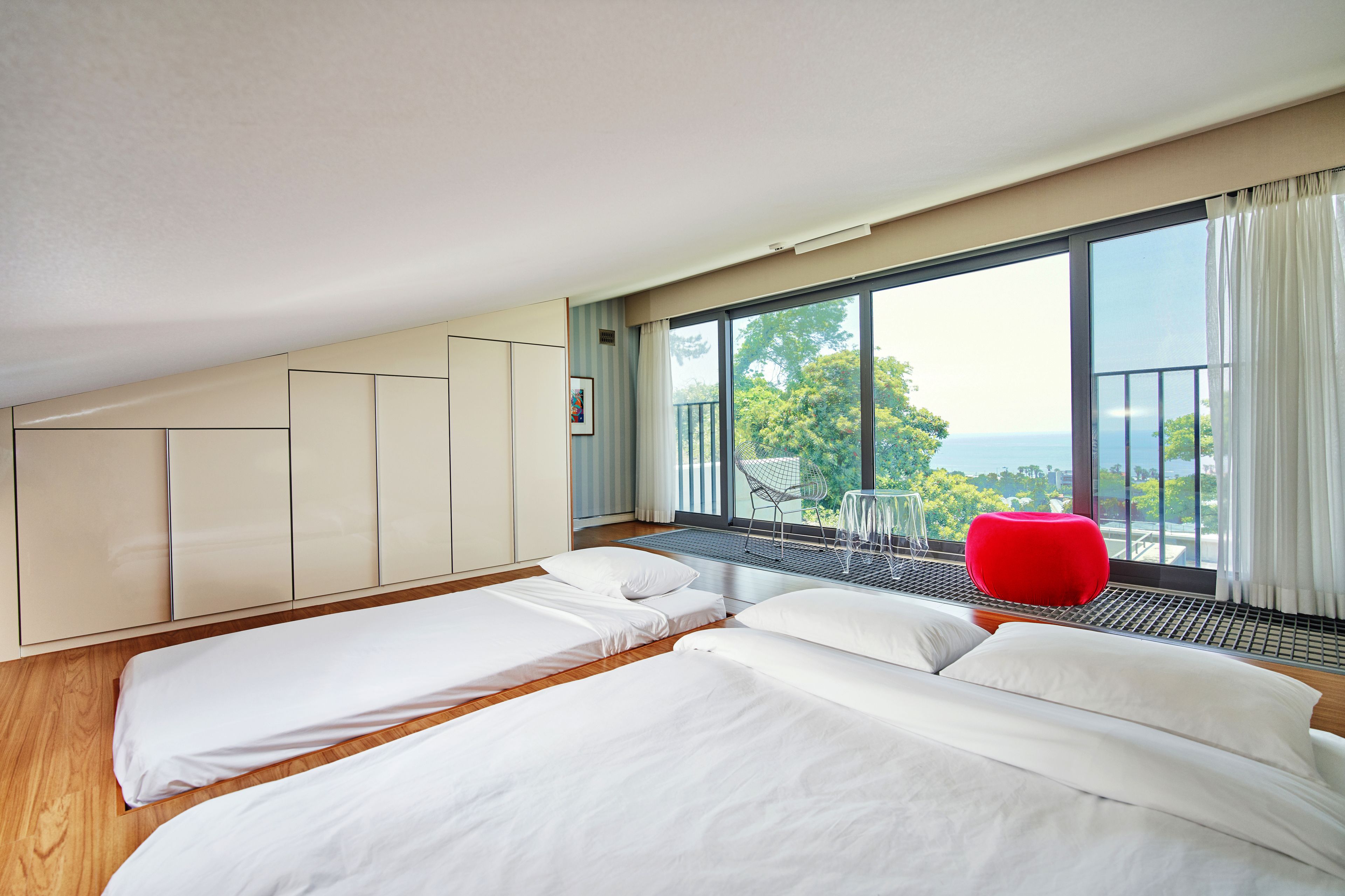 A modern bedroom with two beds, a red chair, and a balcony with a view.