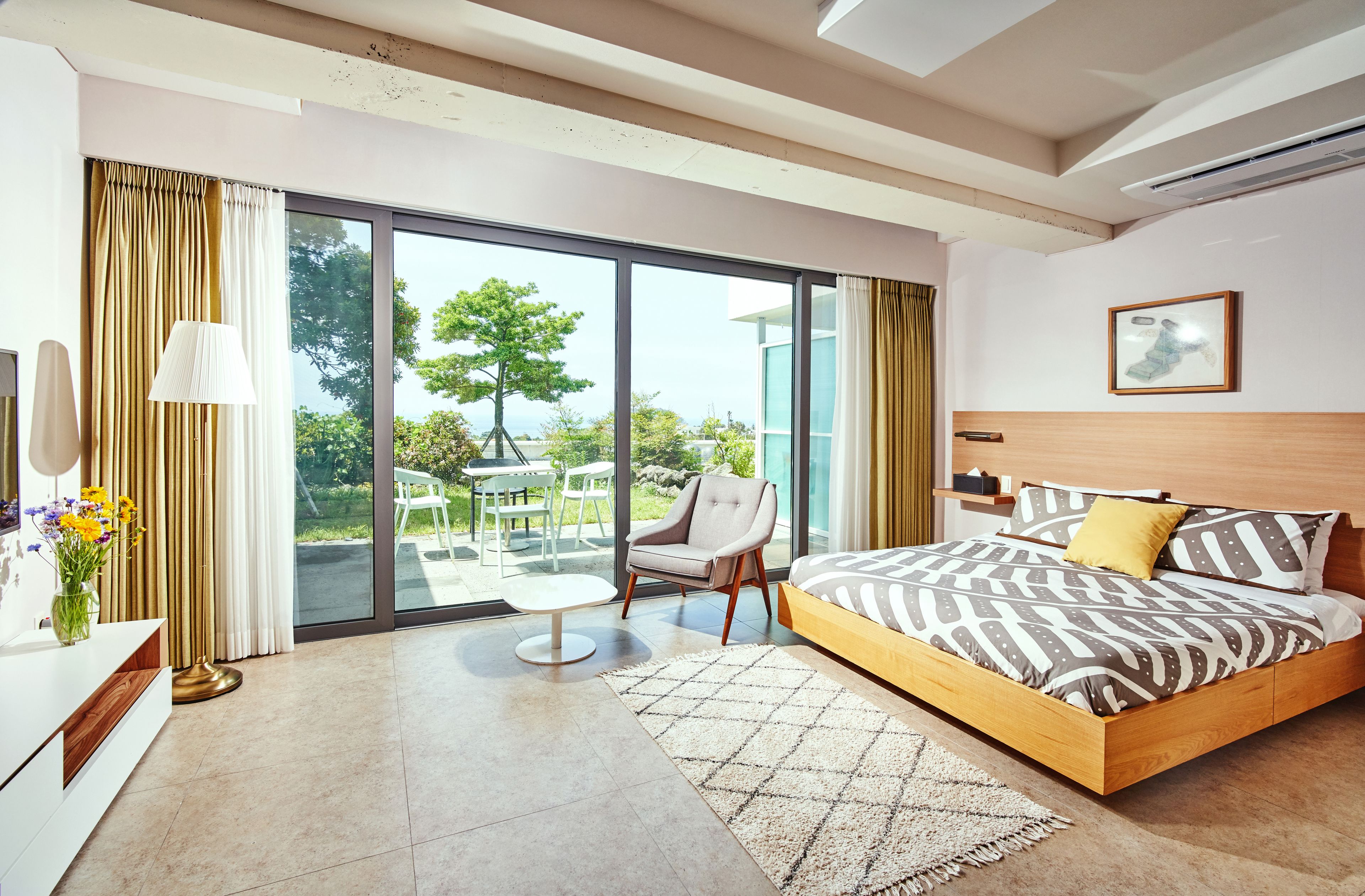 A modern bedroom with a large bed, a chair, a small table, and a view of the outdoors through a sliding door.