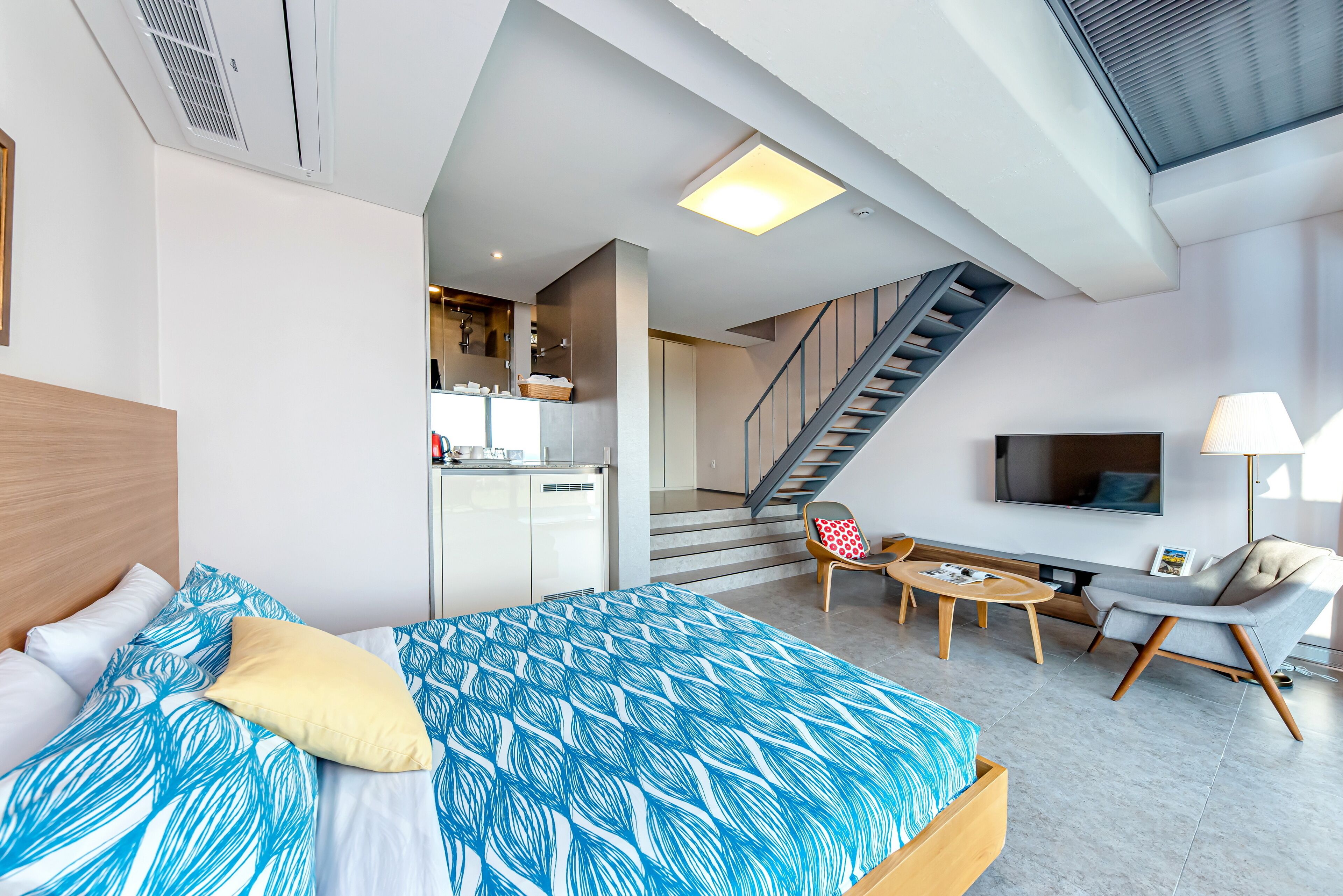 A modern hotel room with a bed, a sofa, a TV, a staircase, and a kitchen area.