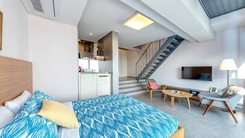 A modern hotel room with a bed, a sofa, a TV, a staircase, and a kitchen area.