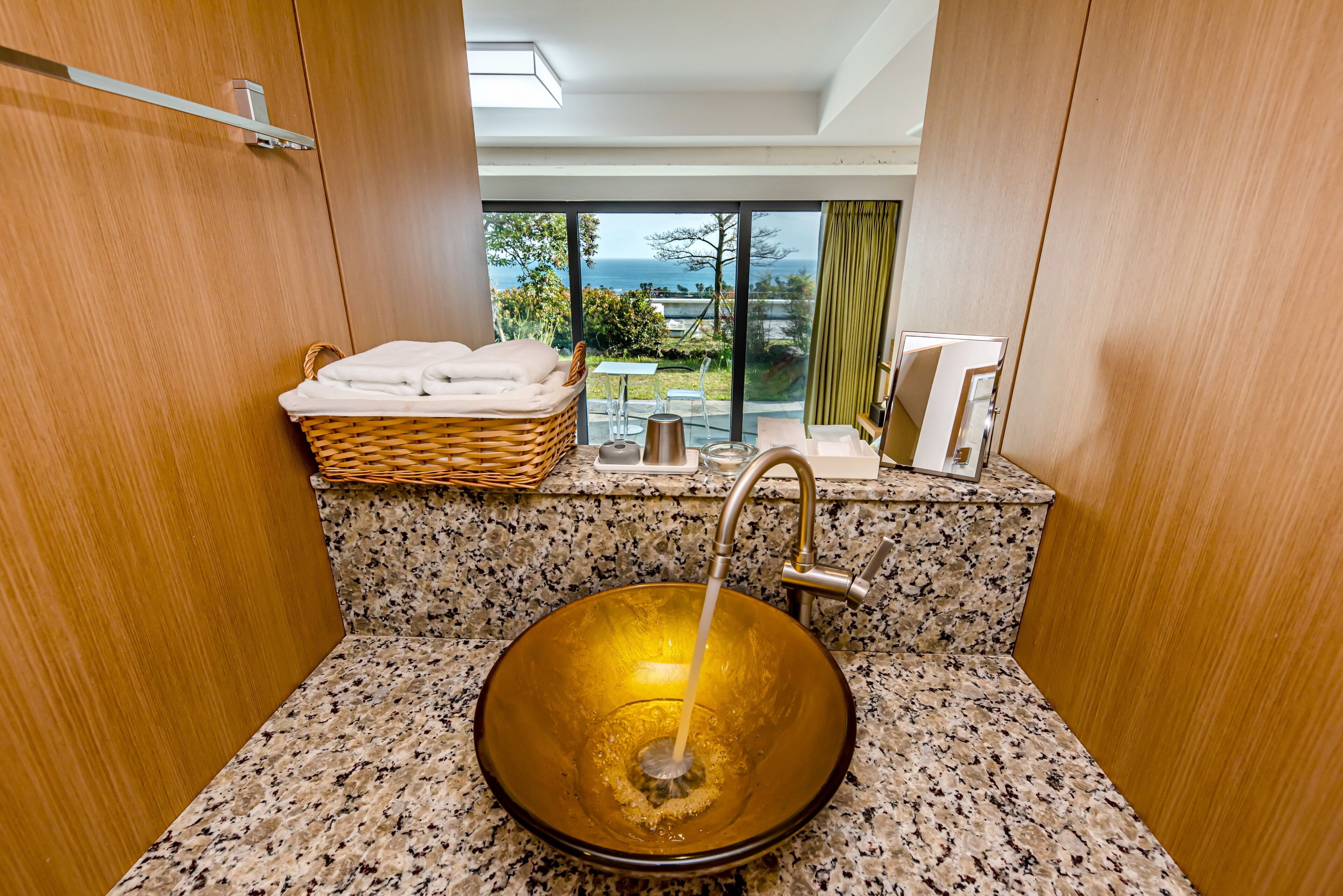 A bathroom with a large, round, amber-colored sink, a wicker basket with towels, and a view of the ocean through large windows.