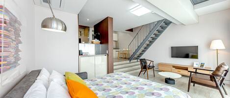 A modern hotel room with a large bed, a desk, a chair, a TV, and a staircase leading to an upper floor.