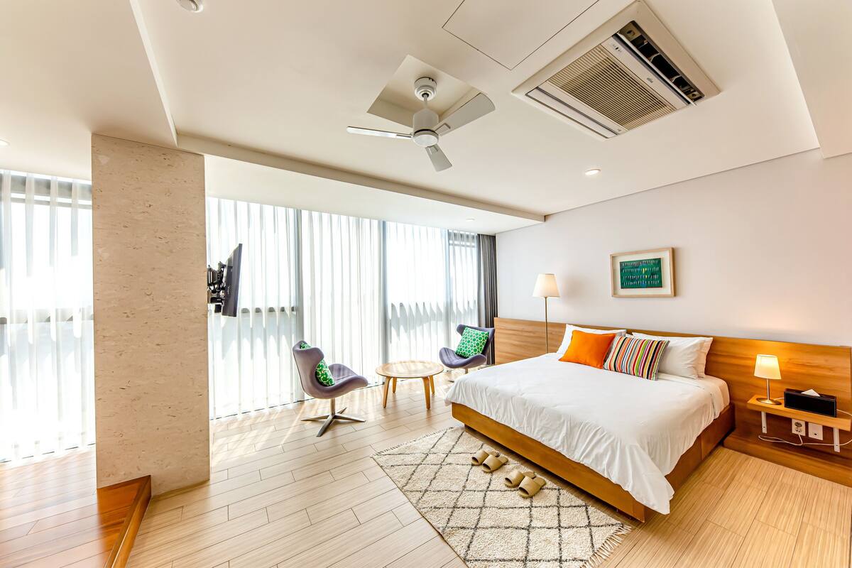 A modern hotel room with a large bed, a ceiling fan, an air conditioning unit, a small round table, two chairs, and a view through large windows.