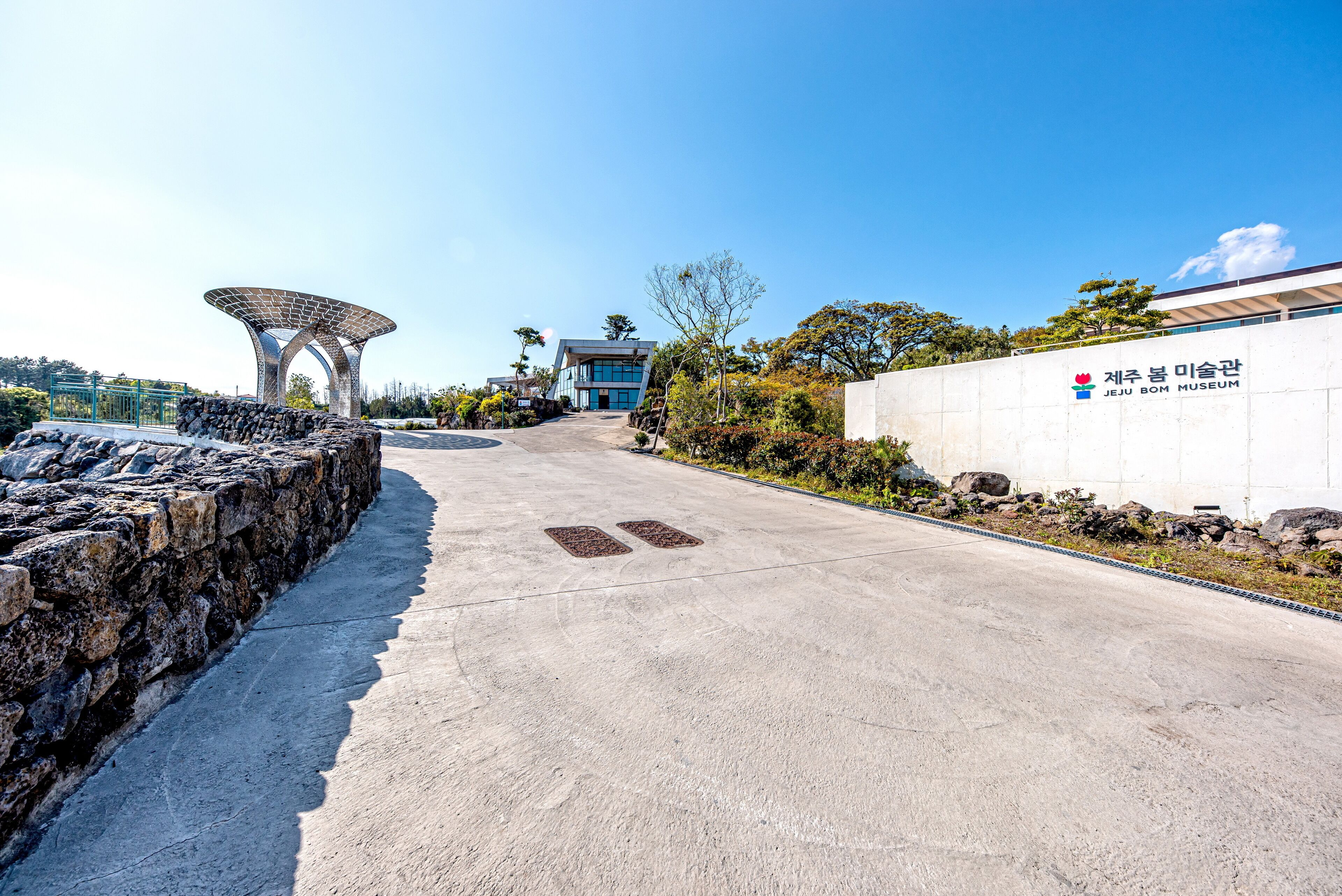 Un camí que condueix a un edifici modern amb una estructura de teulat característica, una paret de pedra a l'esquerra i un rètol per al Jeju Bom Museum.