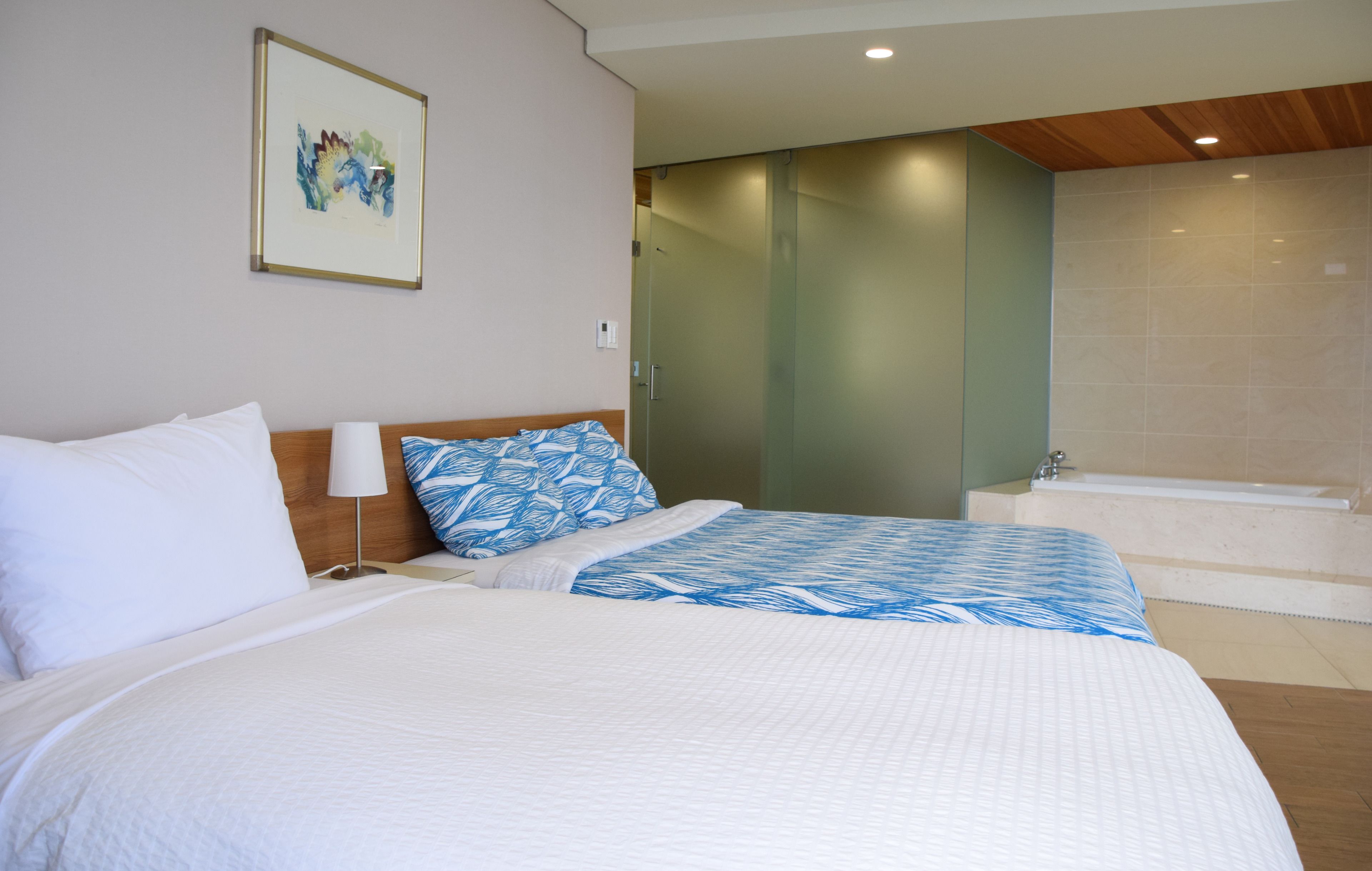 A hotel room with a large bed, a bedside table with a lamp, a framed artwork on the wall, and a bathroom with a bathtub.