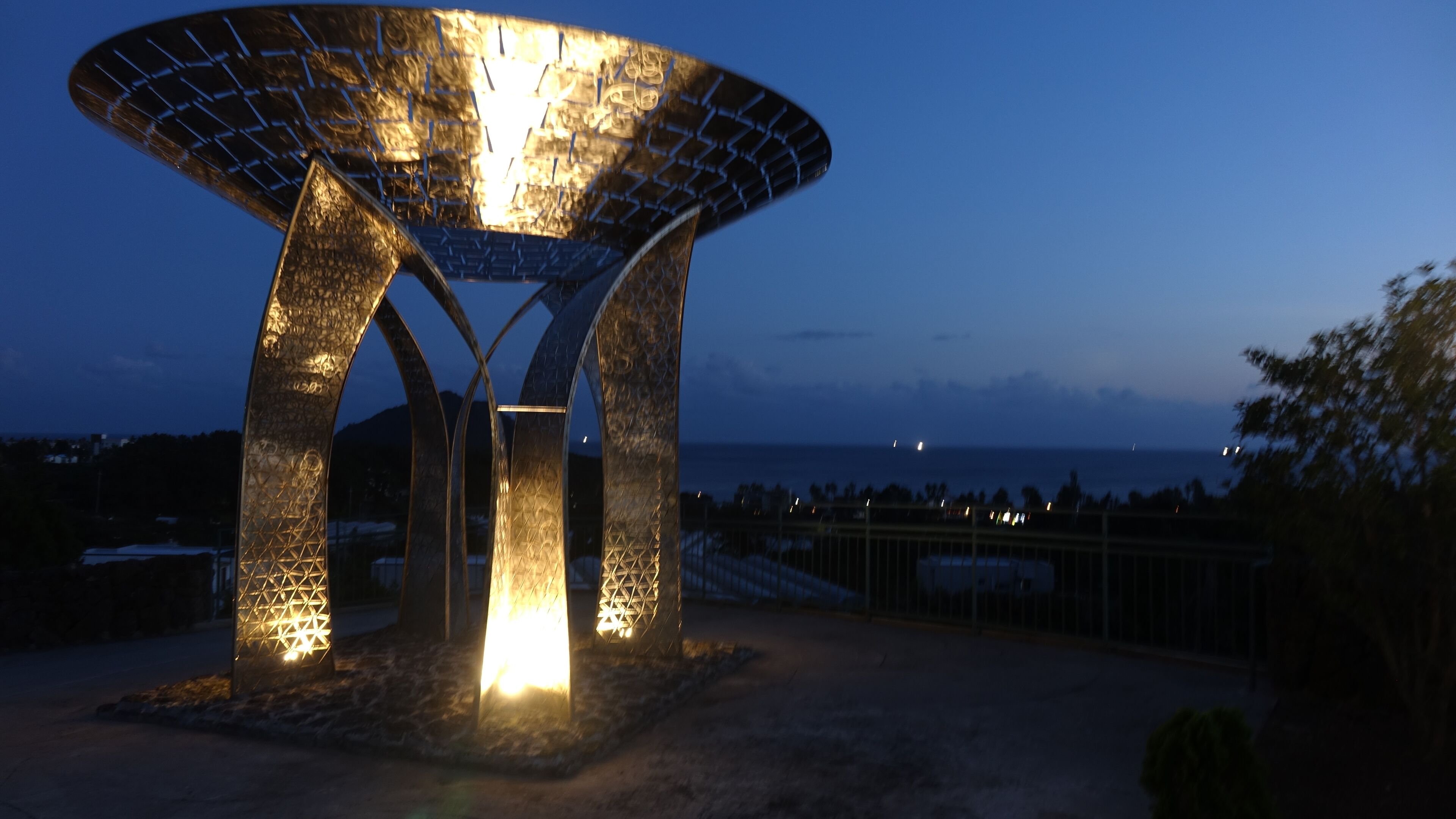 Una escultura metálica con diseño geométrico, iluminada por luces, sobre un cielo crepuscular.