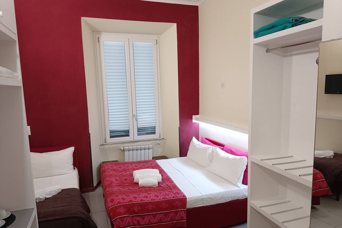 Quarto com cama, radiador, janela com persianas, guarda-roupa e prateleira com toalha.