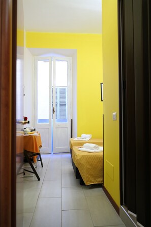 A small, brightly colored room with a yellow wall, a white door, and a bed with a yellow bedspread. There is a small table with a chair and a window with blinds. - Enjoy Roma (Rome)