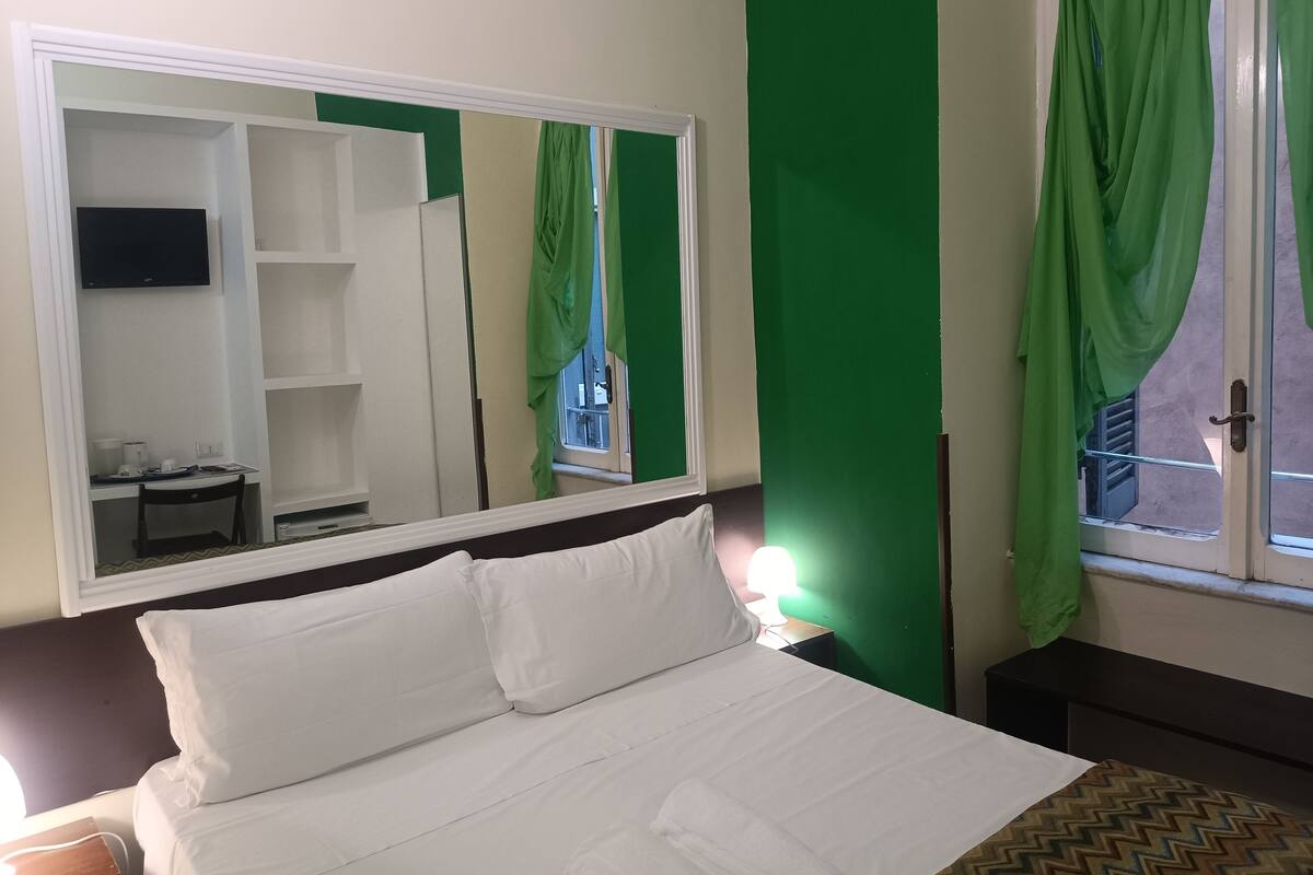 Quarto de hotel com uma cama grande, um guarda-roupa, uma escrivaninha com cadeira e uma TV fixada na parede.