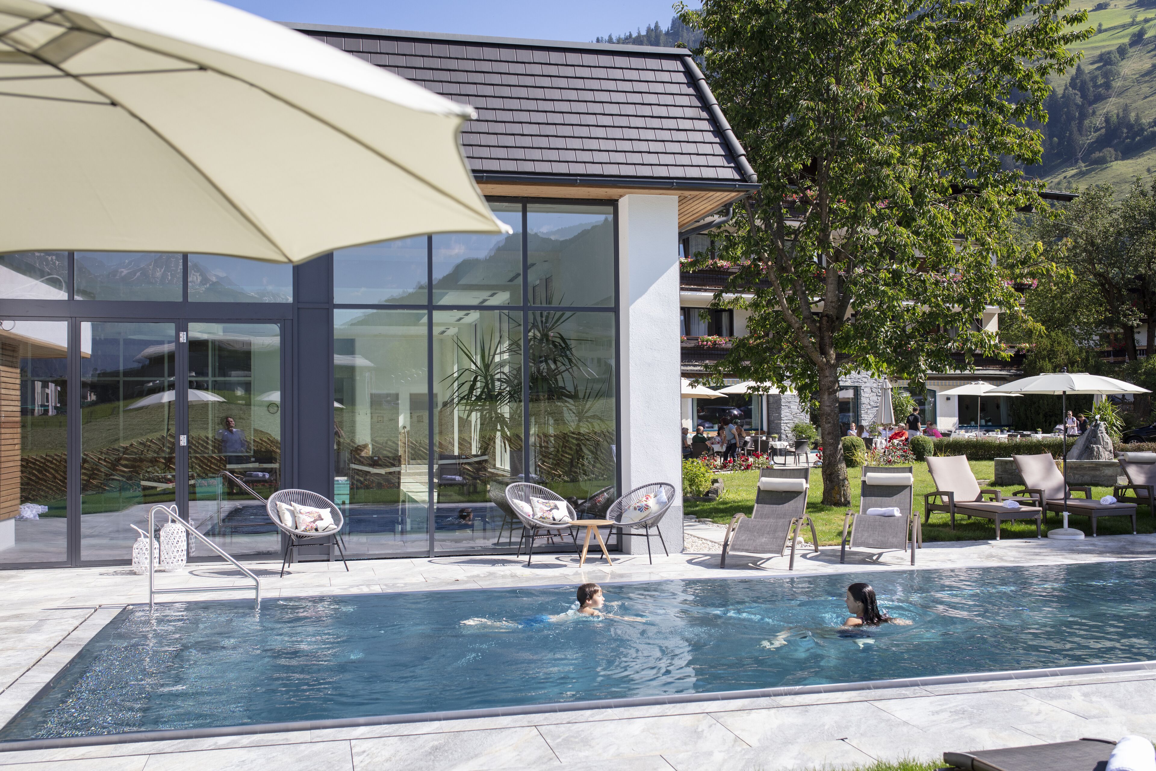 Piscina con sillones de descanso y sombrillas, rodeada por un edificio moderno con grandes ventanales y vistas a montañas.