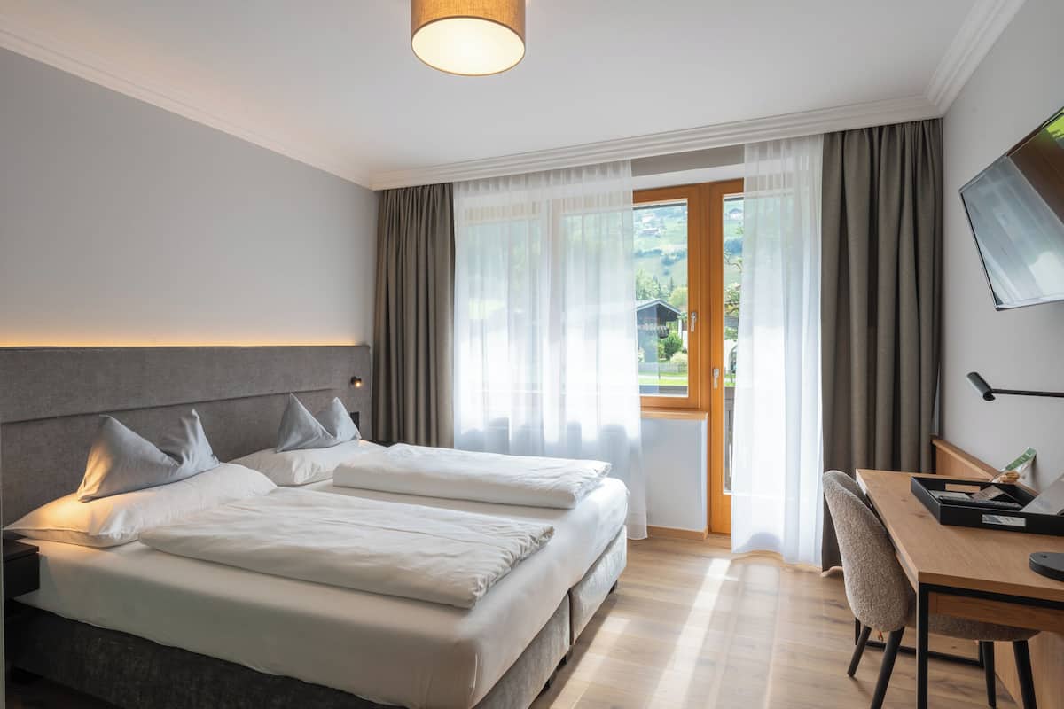 Een hotelkamer met een tweepersoonsbed, een bureau met stoel, een aan de muur bevestigde televisie en uitzicht op groen buiten.