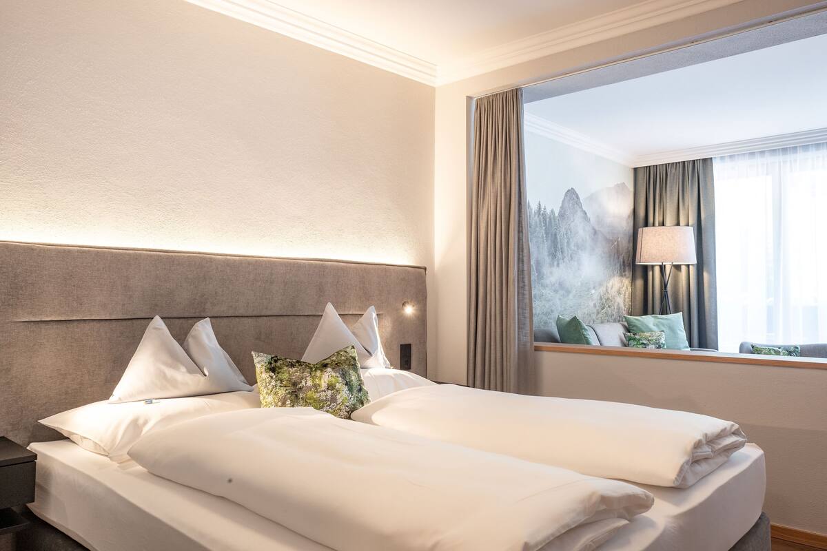 Een hotelkamer met een groot bed, een nachtkastje, een raam met gordijnen en uitzicht op bergen.
