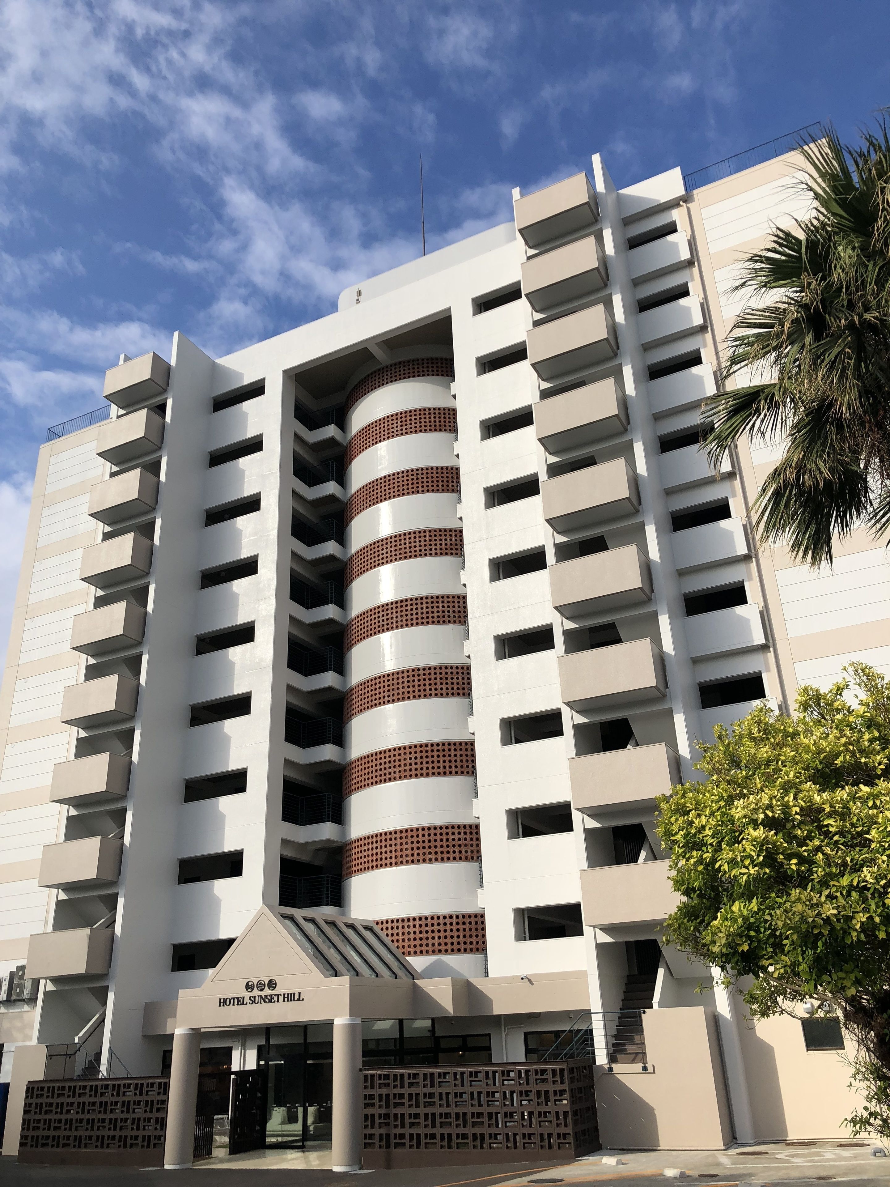 A modern hotel with a central cylindrical tower, multiple balconies, and a clear sky.