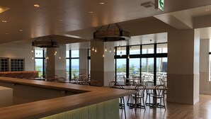 A spacious bar area with a wooden counter and seating, large windows offering a view of the outdoors, and a clean, modern interior design. - Hotel Sunset Hill (Onna)