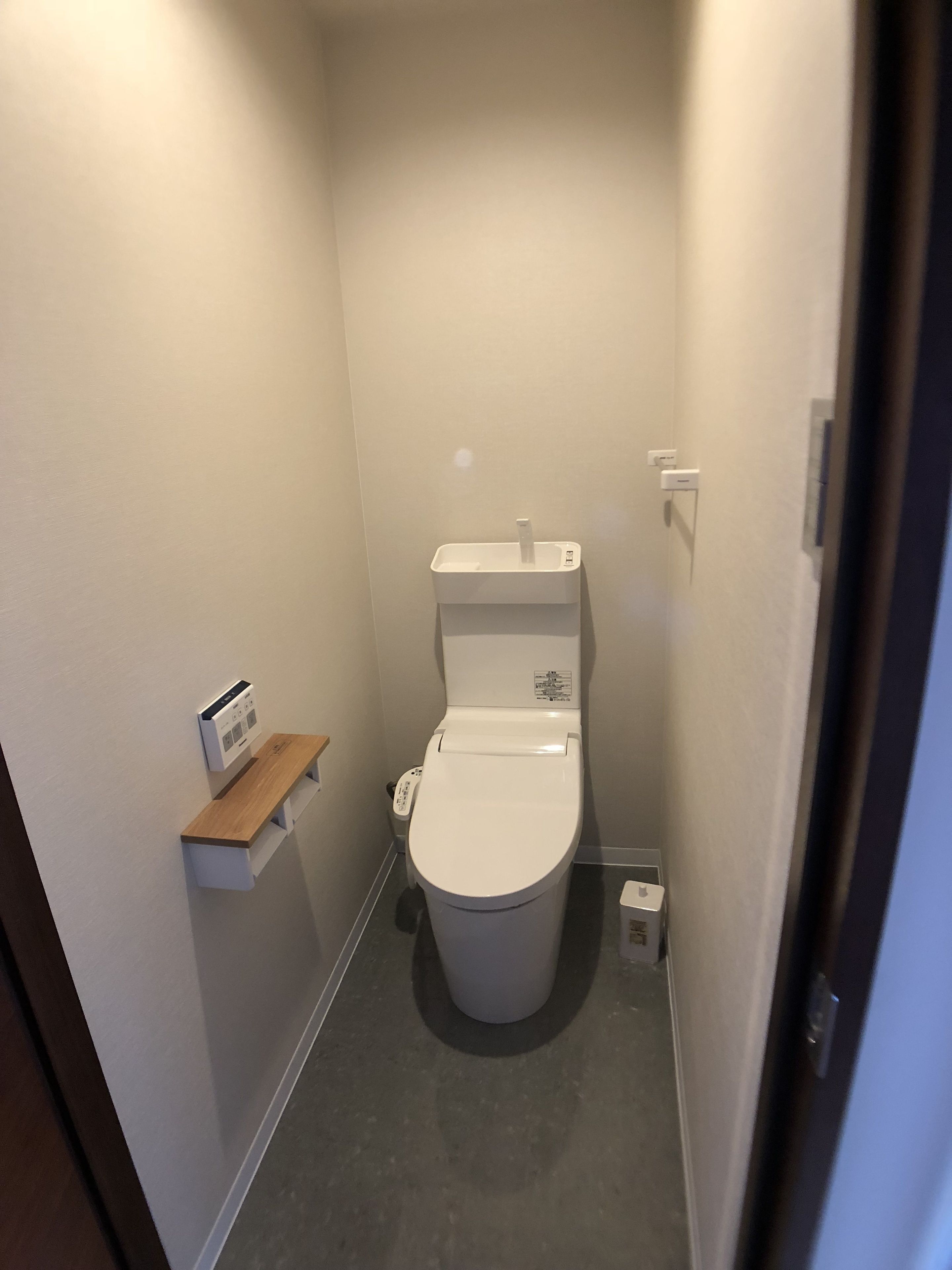 A small, narrow bathroom with a toilet, a wall-mounted shelf, and a small trash bin.