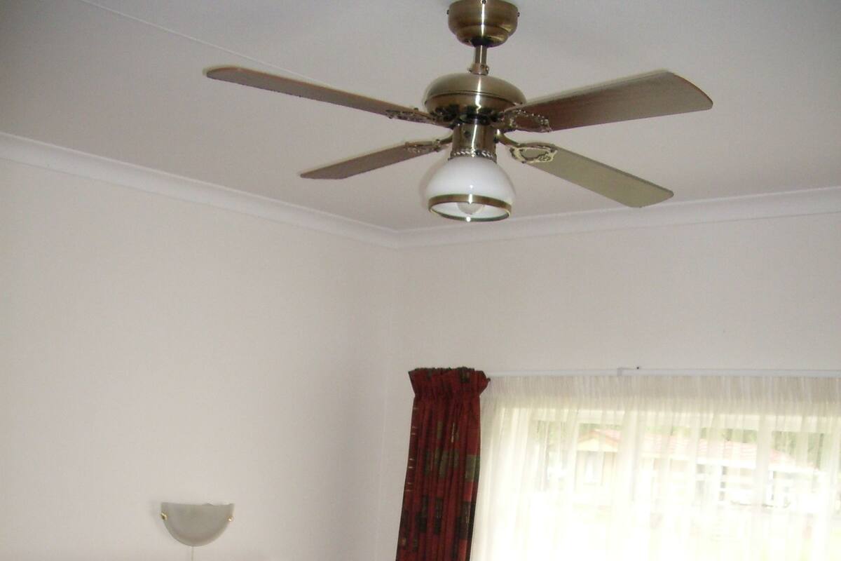 A bedroom with a bed, a nightstand, a wooden headboard, a ceiling fan, and a window with curtains.
