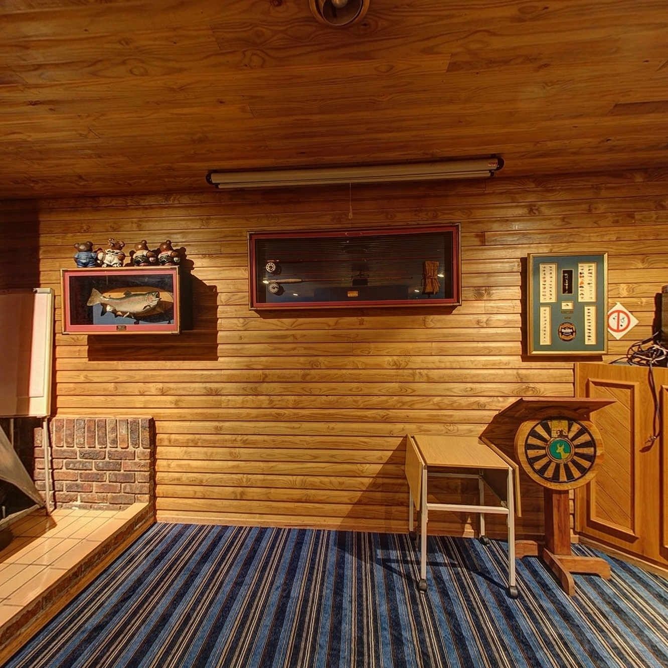 Interior de cabana de madeira com lareira, quadro de peixe emoldurado, relógio e alvo de dardos.