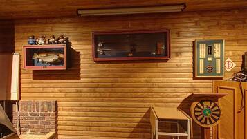 Interior de cabana de madeira com lareira, quadro de peixe emoldurado, relógio e alvo de dardos.