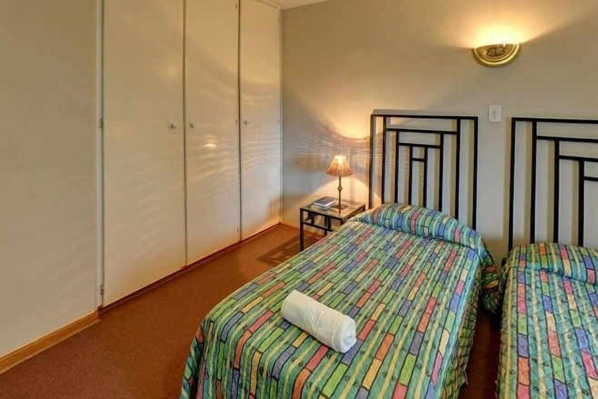 A bedroom with two beds, a nightstand, and a wall-mounted lamp.
