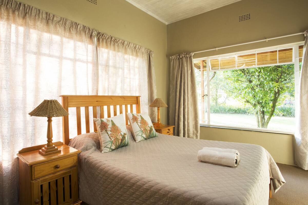 A neatly made bed with a quilted cover, two bedside lamps, a wooden nightstand, and a window with curtains.