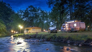 Un camping con una cabaña pequeña y una caravana junto a un río al anochecer.