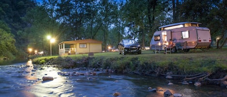Un camping con una cabaña pequeña y una caravana junto a un río al anochecer.