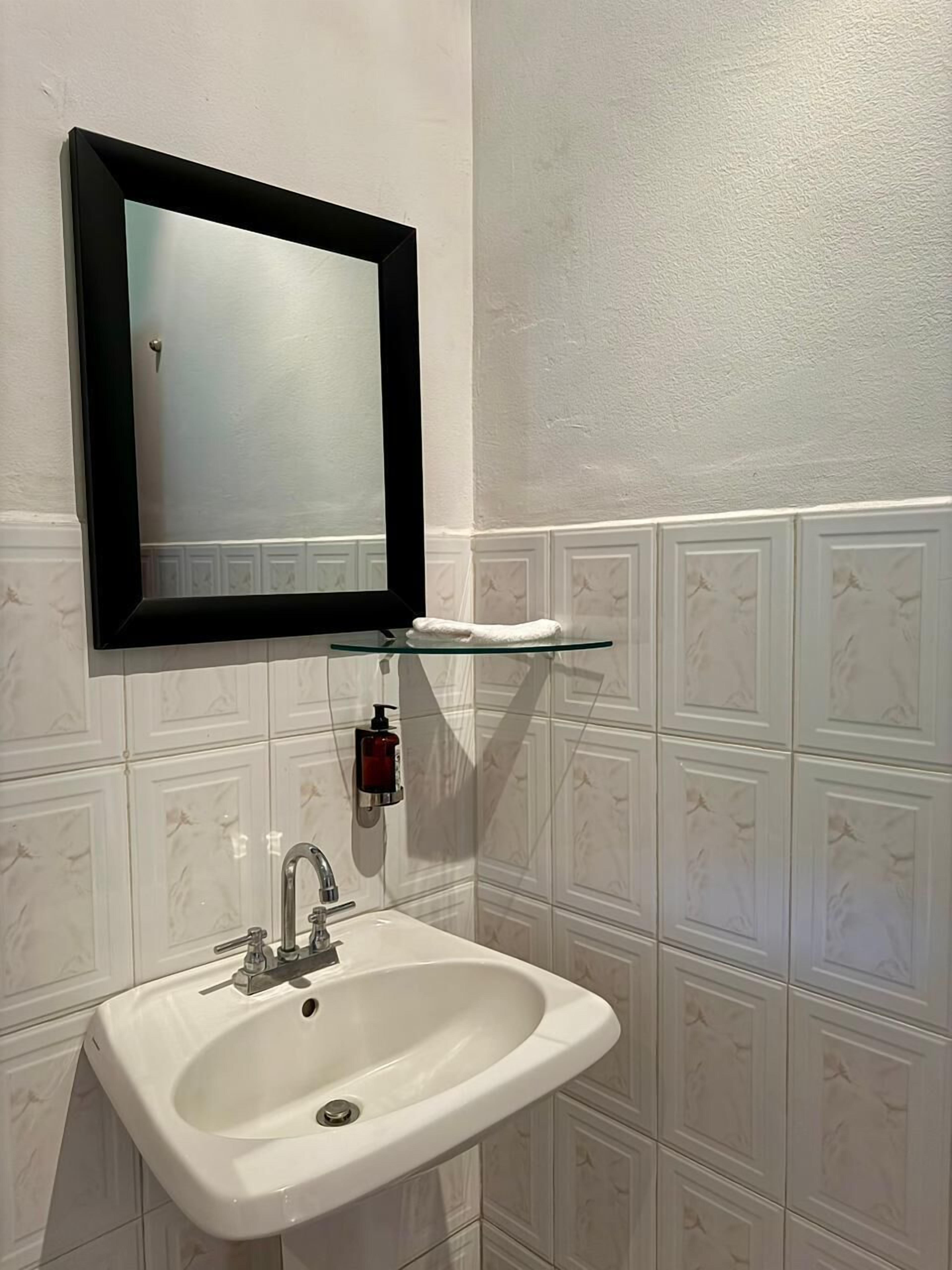 A bathroom with a white sink, a black-framed mirror, and a soap dispenser on a glass shelf.