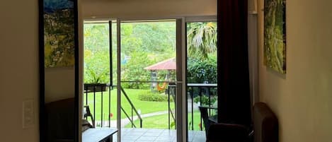 Amplia habitación con un gran ventanal corredero que da a un balcón con vistas a un jardín exuberante y una construcción de tejado rojo.