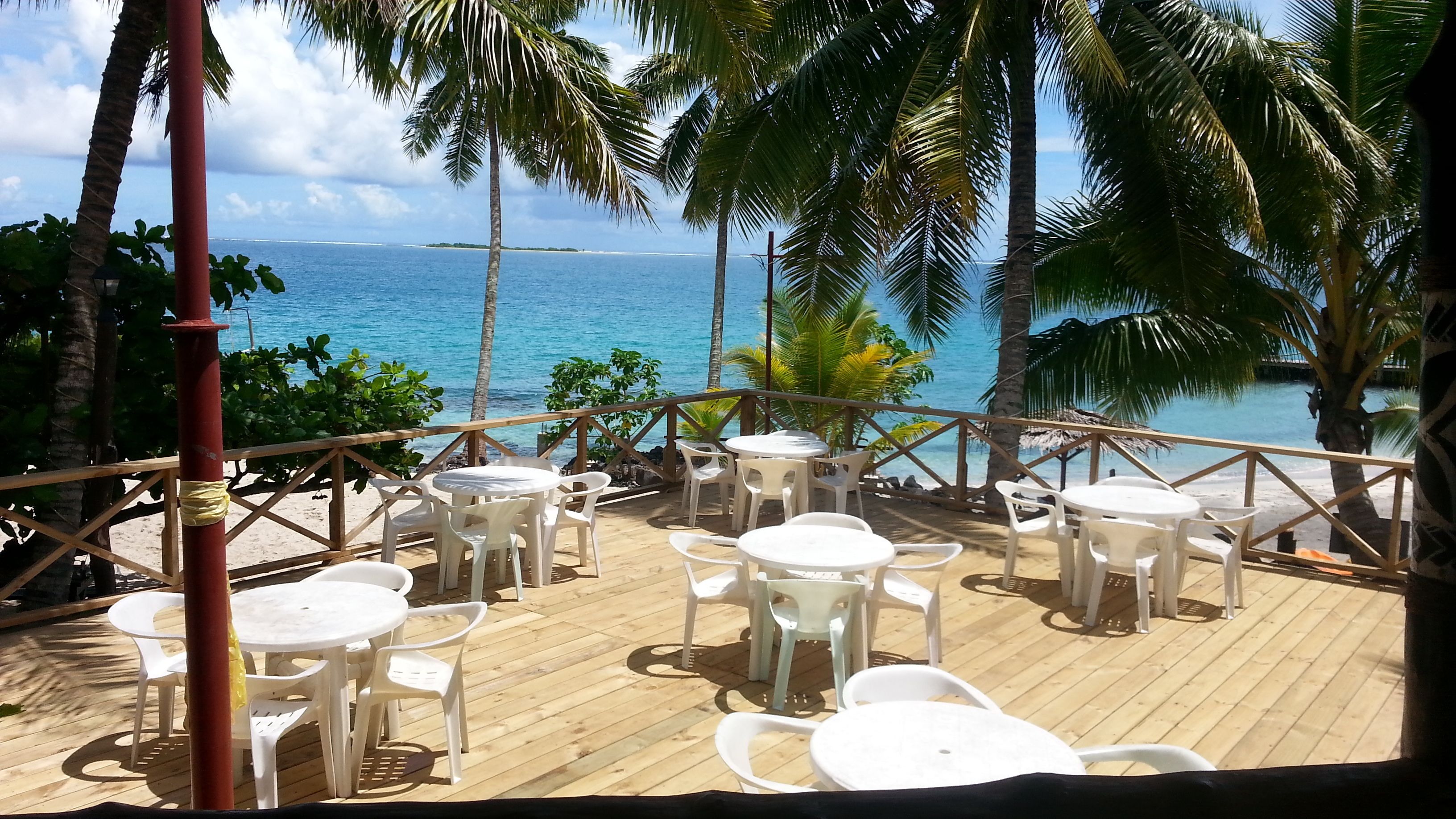 A wooden deck with white plastic tables and chairs, overlooking a beach and the ocean.