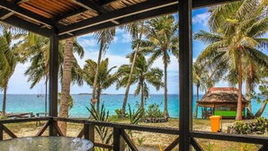 A covered outdoor area with a table and chairs, overlooking a beach with palm trees and a thatched-roof structure. - Va-i-moana Seaside Lodge (Asau)