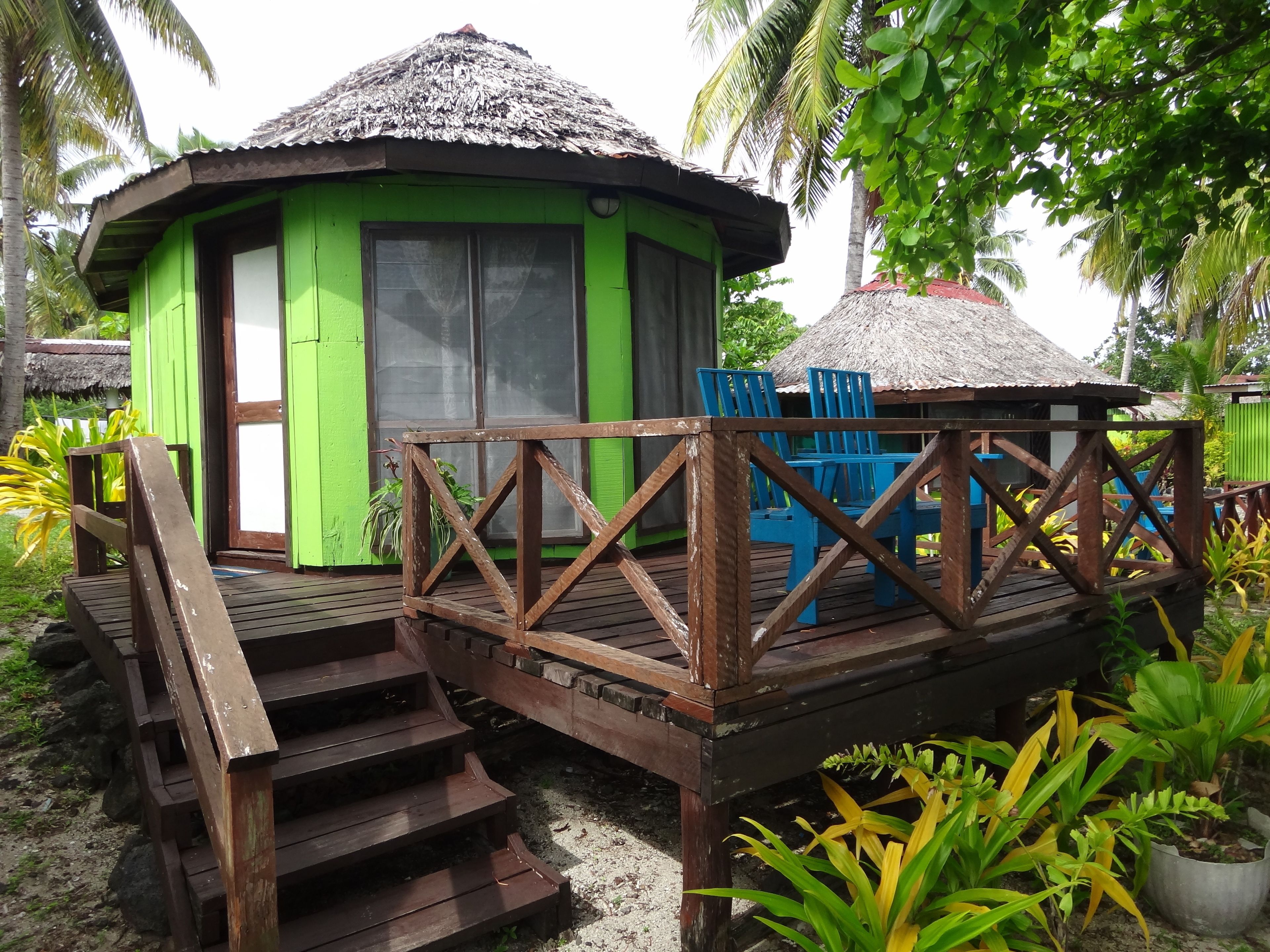 A small, green building with a thatched roof, wooden deck, and blue chairs.